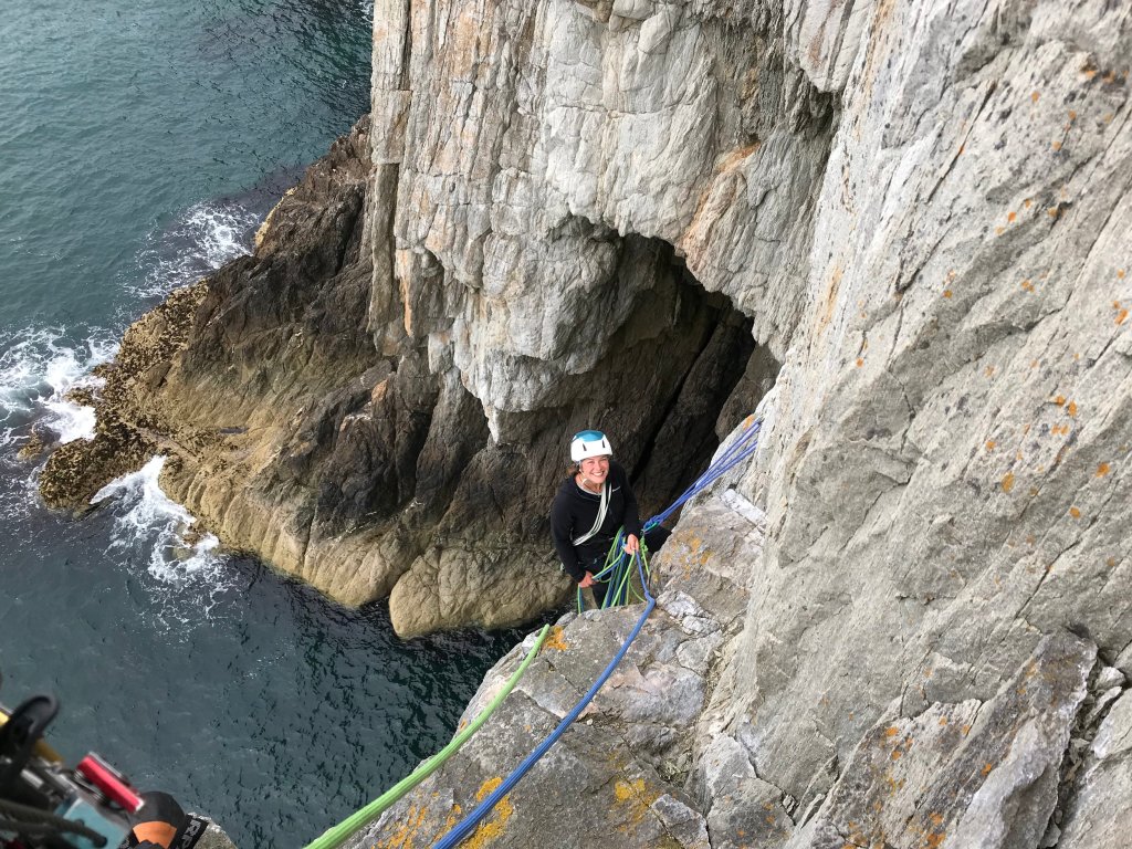 smiling female climber on a hanging belay to the side of a coastal lawn with the sea below